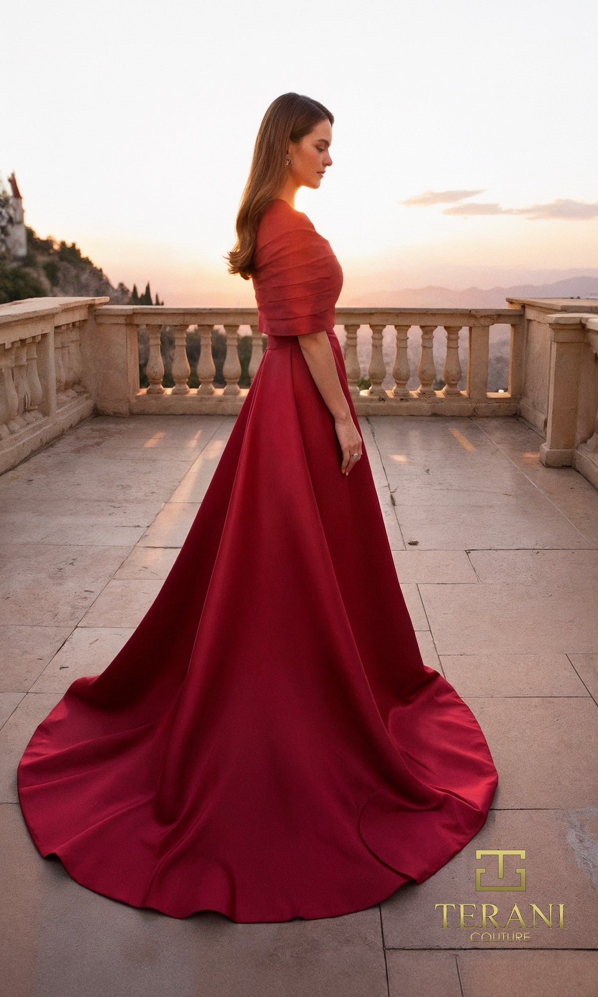 A woman in Terani 261M6415, a red matte satin floor-length mother-of-the-bride gown with a fitted bodice and long train, stands sideways on a stone terrace at sunset. The